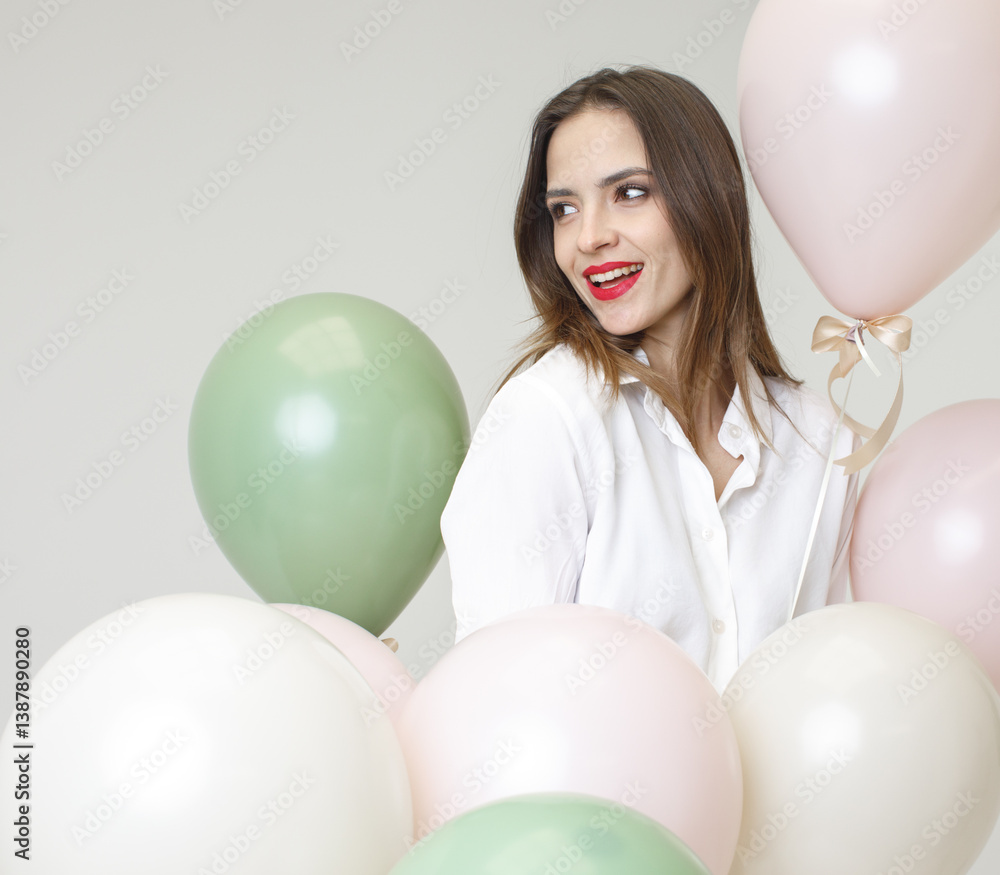 Happy young woman with red lipstick smiling, looking left side among pastel balloons. Light background, festive mood, cheerful and bright atmosphere.