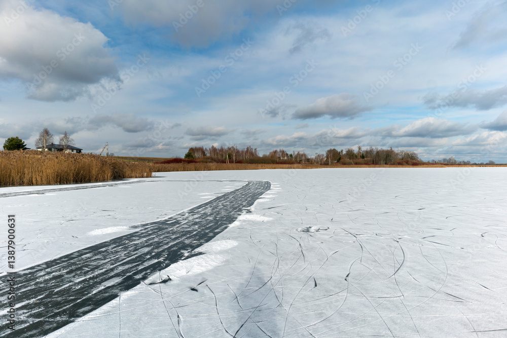 Fototapeta premium Frozen Lake with Cracked Ice and Winter Landscape. Scenic winter landscape featuring a frozen lake with a prominent crack or ice path cutting through the snow-covered surface. In the distance, dry ree