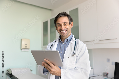Happy man therapist dressed in white uniform using modern digital tablet device working online, consulting clinic patients remotely, standing alone in contemporary medical office. Technology, workflow