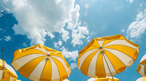 Yellow and white umbrellas viewed from below, set against a sunny blue sky filled with clouds. Concept of summer relaxation outdoors