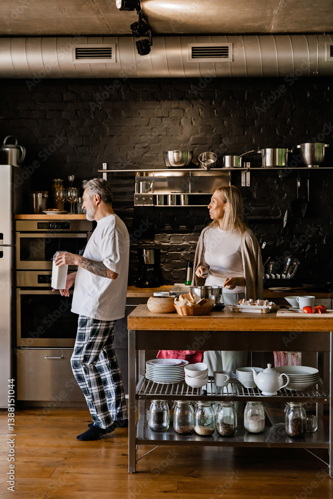 A woman stands at a table holding a whisk while looking at a man walking next to her holding a carton of milk, in the kitchen, before retirement A woman stands at a table holding a whisk while looking at a man walking next to her holding a carton of milk, in the kitchen, before retirement