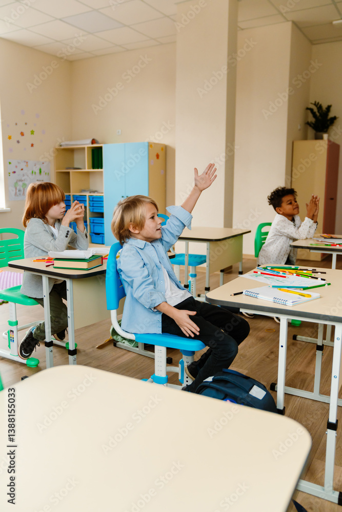 Obraz premium A group of three schoolchildren sit at desks and look ahead while one of them raises his hand, at school, in the classroom