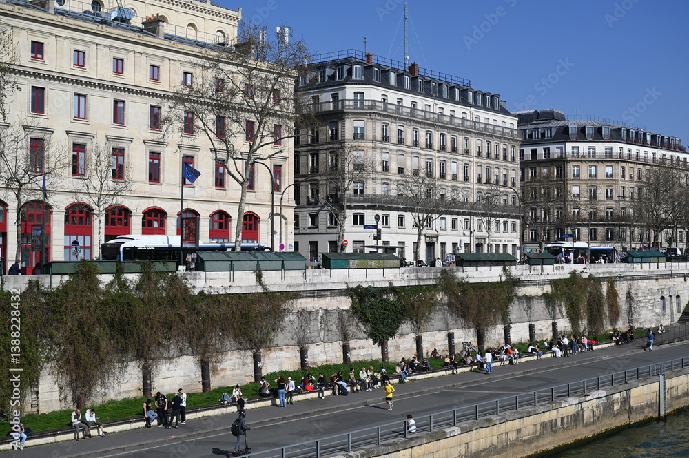 Naklejka premium Seine river embankment in Paris with classic Haussmann buildings, bridge, and blue sky. Scenic cityscape of the French capital with historical architecture and calm water.