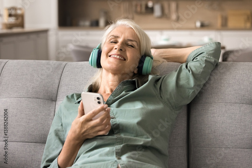 Enjoying music. Portrait smiling mature woman wear digital headphones recline on sofa listen to songs from chosen playlist with closed eyes chill relax catching meditative sounds online on cellphone