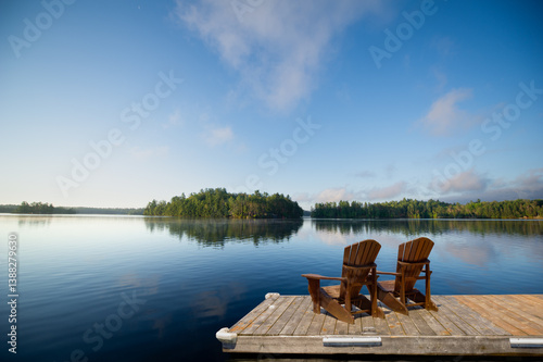 Two Adirondack chairs sit on a wooden dock overlooking a calm Muskoka lake, with fluffy clouds and lush green trees mirrored in the still water. Space for copy.