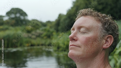 A middle-aged man with curly hair stands by a quiet lake with his eyes closed, enjoying a moment of peace and mindfulness in nature.