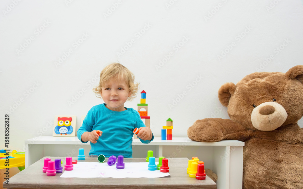 Cute little child boy having fun at home playing with colorful stamps on paper. Educational indoor activity for kids.