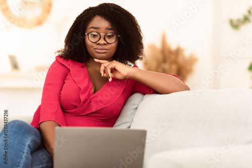 Quadro su tela Portrait of confused skeptic black curly woman in eyeglasses looking at her lapt