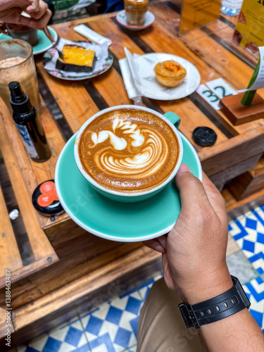 Obraz na płótnie Magic Coffee with two ristretto shots and steamed milk served in a turquoise cup, held over a wooden table with pastries and drinks, capturing a cozy café moment. w salonie