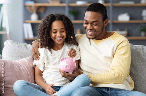 Foto Black girl and father putting coin into piggy bank, sitting on sofa at home