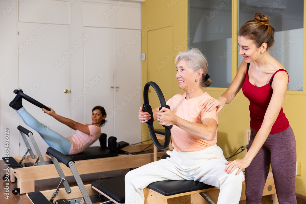 Mature European woman who cares about her health is engaged in Pilates, performing an exercise with a special ring, where ..the coach helps to do it correctly