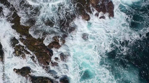 Powerful ocean waves crash over jagged coastal rocks, creating frothy white foam against the deep blue sea. Aerial view captures the force of nature in motion.