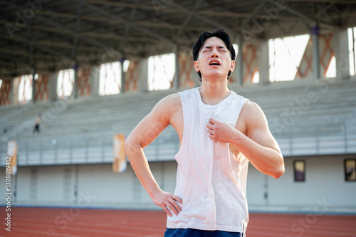 Asian man is standing with one hand on waist and other hand grabbing a white tank top. Hot or tired.