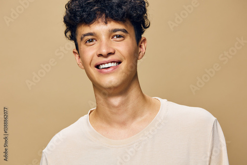 Obraz na płótnie Smiling young man with curly hair wearing a light shirt, set against a warm, beige background that enhances the cheerful and friendly atmosphere of the portrait w salonie