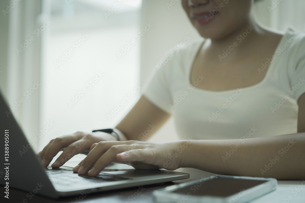 A woman is typing on a laptop computer while smiling. She is wearing a white shirt and has a watch on her wrist. There are two cell phones on the table, one on the right and one on the left A woman is typing on a laptop computer while smiling. She is wearing a white shirt and has a watch on her wrist. There are two cell phones on the table, one on the right and one on the left