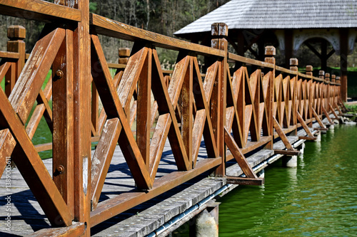 pedestrian wood footbridge over green lake with gazebo. parks and outdoors. cross braced balustrade with posts. brown stained finish. perspective view. soft green background. wood planks on beams