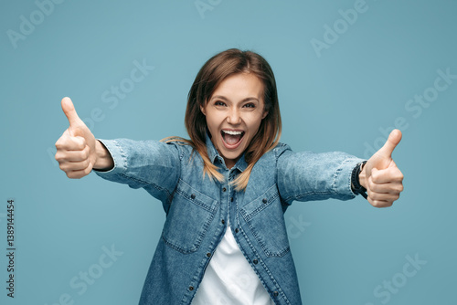 Cheerful young woman in jeans shirt showing thumbs up over blue background. Look in camera
