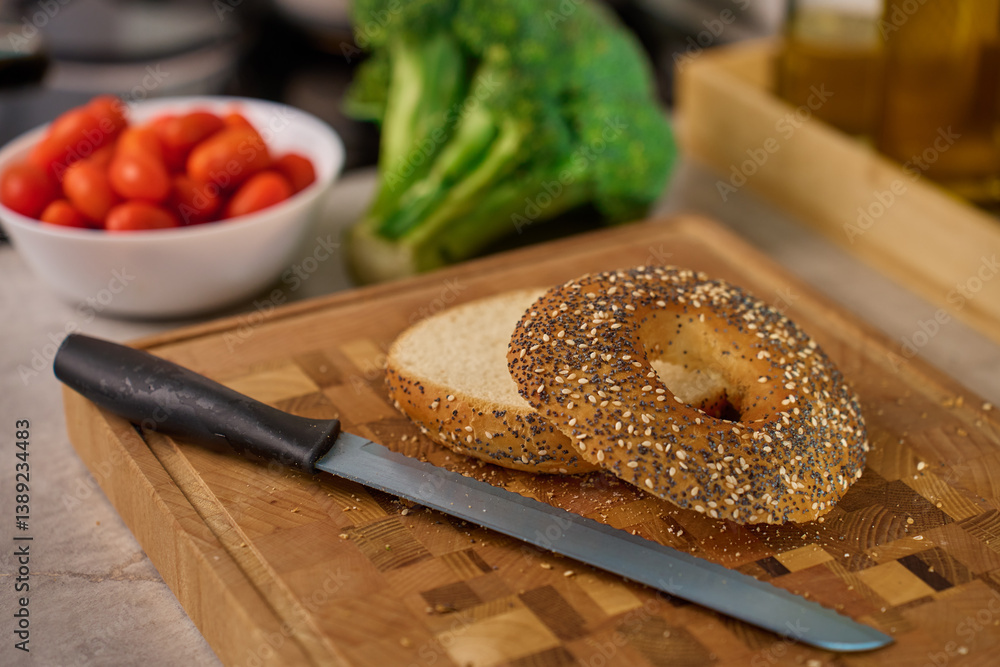 Sliced bagel on wooden cutting board with cherry tomatoes, broccoli and kitchen tools in background. Preparation of healthy breakfast or snack in home kitchen