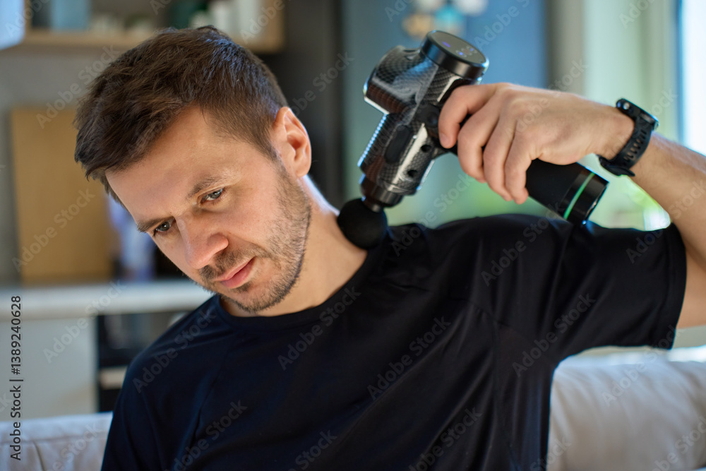 Man applies massage gun to relieve tension in neck and shoulder area. Male athlete doing self-massage therapy with percussion device for recovery after training