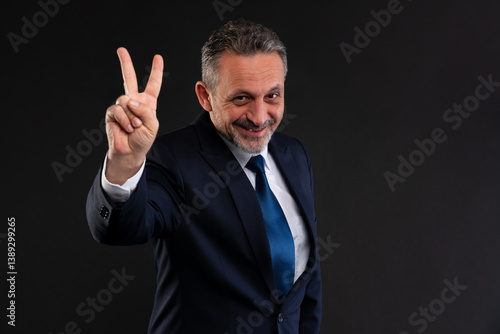 Confident mature man in formal suit smiling and holding up two fingers in a peace or victory gesture, standing against a dark background.
