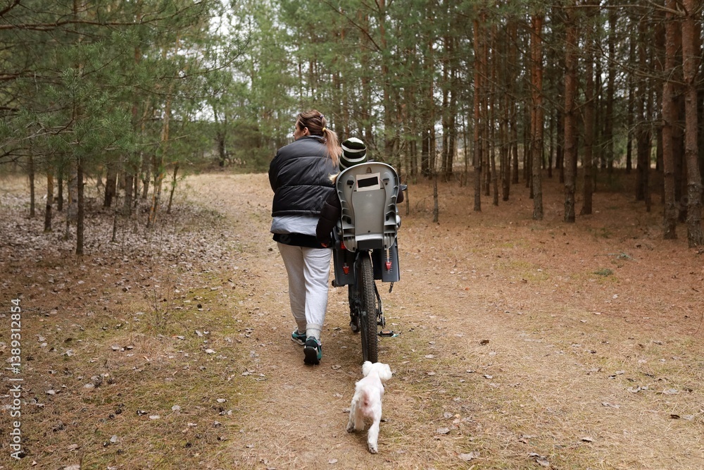 Woman walks with child on bicycle in forest while dog runs alongside them during a sunny afternoon outing