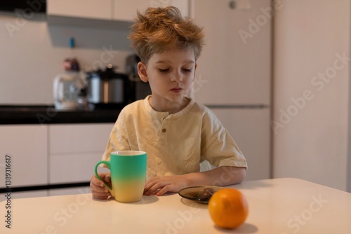 Obraz na płótnie Young boy enjoying breakfast with a cup and fruit in a bright kitchen during morning hours w salonie