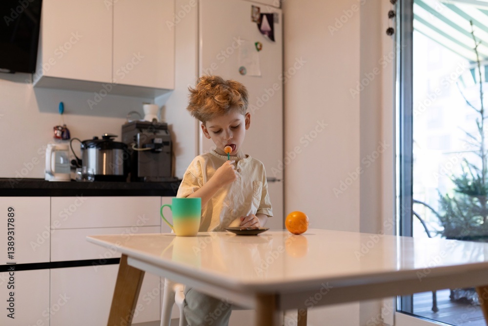 Child enjoying a colorful lollipop while sitting at a modern kitchen table with fruit and a beverage in the morning