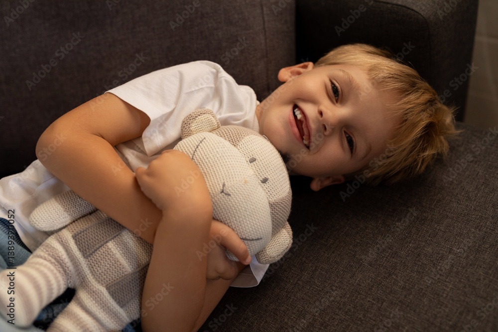 Boy smiles while hugging a soft toy on a cozy couch in a living room during the afternoon