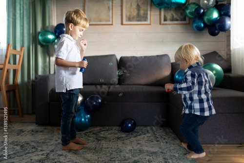 Two young boys playing with bubbles in a festive room decorated with balloons and a cozy couch in the background