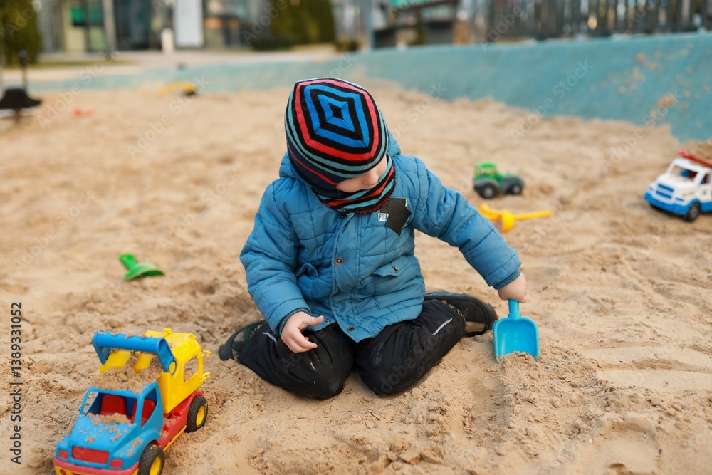Young child plays in a sandbox with colorful toy trucks on a sunny day at a playground