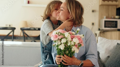 Young boy gives his mother a bouquet of flowers at home in a warm and cozy setting. She smiles with joy and gratitude. Concept of Mother's Day celebration and family love. Mid-range shot natural light