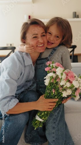 Young boy gives his mother a bouquet of flowers at home in a warm and cozy setting. She smiles with joy and gratitude. Concept of Mother's Day celebration and family love. Mid-range shot natural light