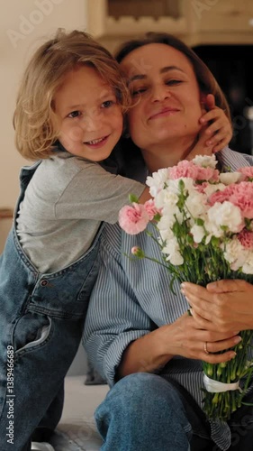 Young boy gives his mother a bouquet of flowers at home in a warm and cozy setting. She smiles with joy and gratitude. Concept of Mother's Day celebration and family love. Mid-range shot natural light