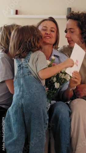 Three children celebrate Mother's Day by giving their mom flowers and a greeting card at home. She smiles, surrounded by love and joy. Concept of motherhood and family connection. Mid-range shot
