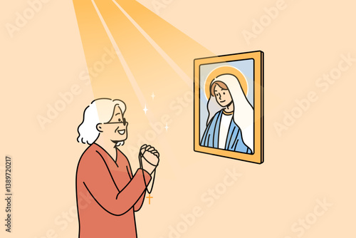 Elderly woman prays to god standing near icon of virgin mary, under rays of light. Gray-haired lady prays holding catholic crucifix in hands and smiling, thanking mother of jesus for life