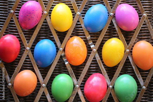 The Easter colorful eggs are lying on the woven table. The eggs are lying in the cells of a metal trivet.