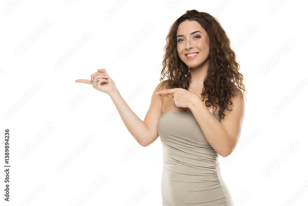 Beautiful young woman with curly hair wearing beige dress pointing with index finger, gesturing to product placement area, white background Beautiful young woman with curly hair wearing beige dress pointing with index finger, gesturing to product placement area, white background