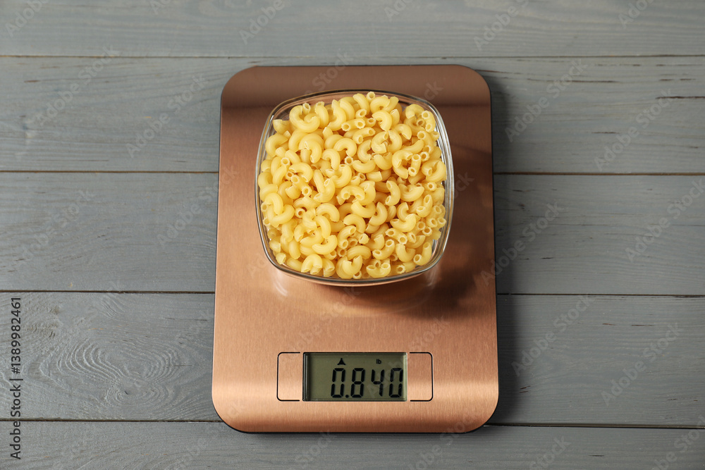 Modern electronic kitchen scale with bowl of raw pasta on grey wooden table, top view