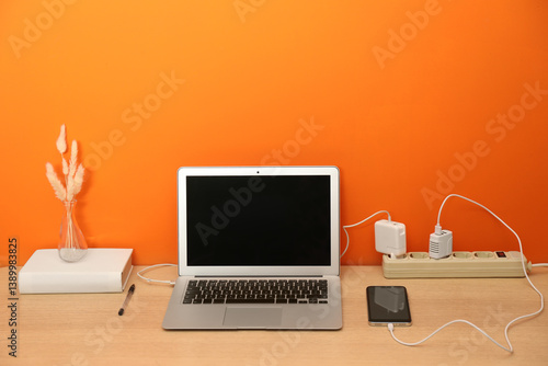 USB adapters with cables plugged into power strip charging laptop and smartphone on wooden table near orange wall