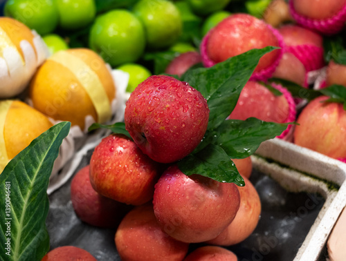 Fresh red apples with dew drops and green leaves, surrounded by vibrant fruits in a market display.