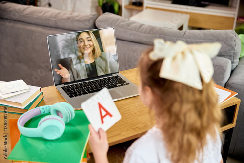 Little girl holding flashcard with letter A, communicating with teacher via laptop, during online alphabet lesson at home. Concept of childhood, online education, remote studying
