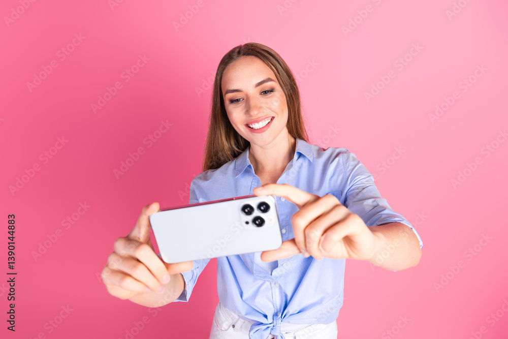 Happy young woman taking a selfie with smartphone against vibrant pink backdrop, wearing trendy blue shirt and enjoying leisure time Happy young woman taking a selfie with smartphone against vibrant pink backdrop, wearing trendy blue shirt and enjoying leisure time