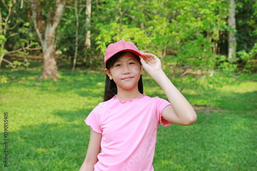 Portrait of smiling Asian girl preteen wearing pink cap hat with hands holding her hat and looking straight at camera while standing in green park.
