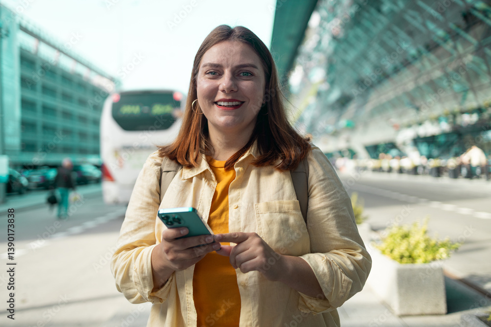 A smiling woman in a yellow shirt and light jacket stands at the airport holding a smartphone. She exudes positive energy and confidence while engaging with her phone in the busy travel environment.
