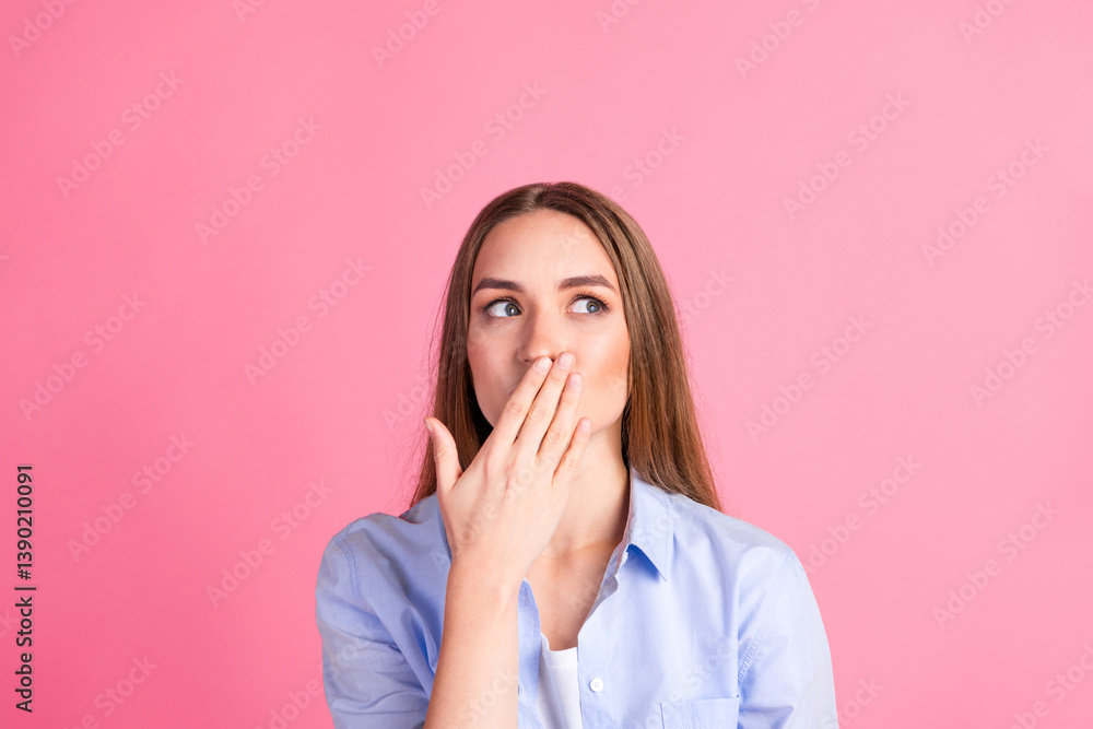 Attractive young woman covering mouth with hand in surprise against pastel pink background Attractive young woman covering mouth with hand in surprise against pastel pink background