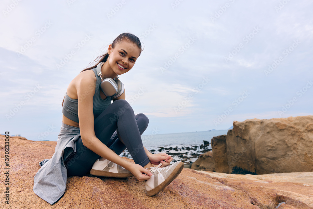Young woman in activewear enjoying nature, tying shoes on rocky beach with headphones, promoting fitness and outdoor lifestyle