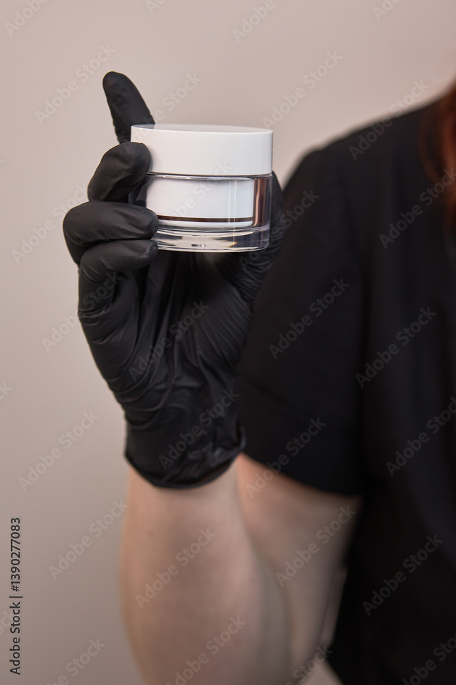 Hand in black glove holding white cosmetic jar, closeup of beauty product in professional care setting, skincare focus concept