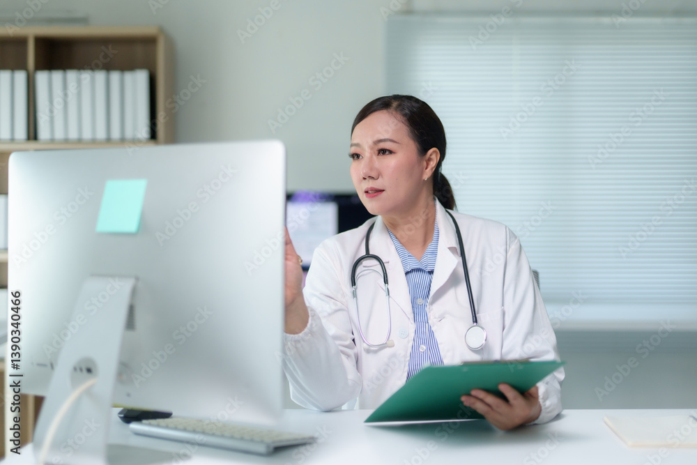 Asian female doctor wearing lab coat and stethoscope having video call with patient using desktop computer in hospital office Asian female doctor wearing lab coat and stethoscope having video call with patient using desktop computer in hospital office