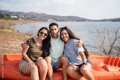 Three latin american friends sitting on an orange kayak by a lake with mountains in the background