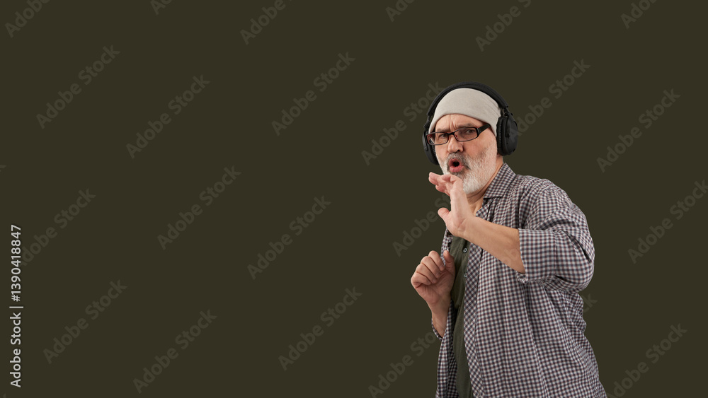An adult gray-haired and gray-bearded man listening to music on headphones. The background is dark green . Copy space.